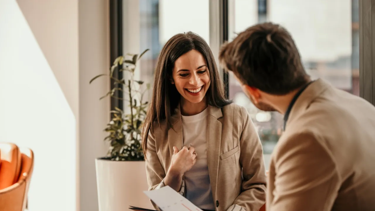 A woman smiles during a business meeting while discussing a report with a colleague in a modern office setting.