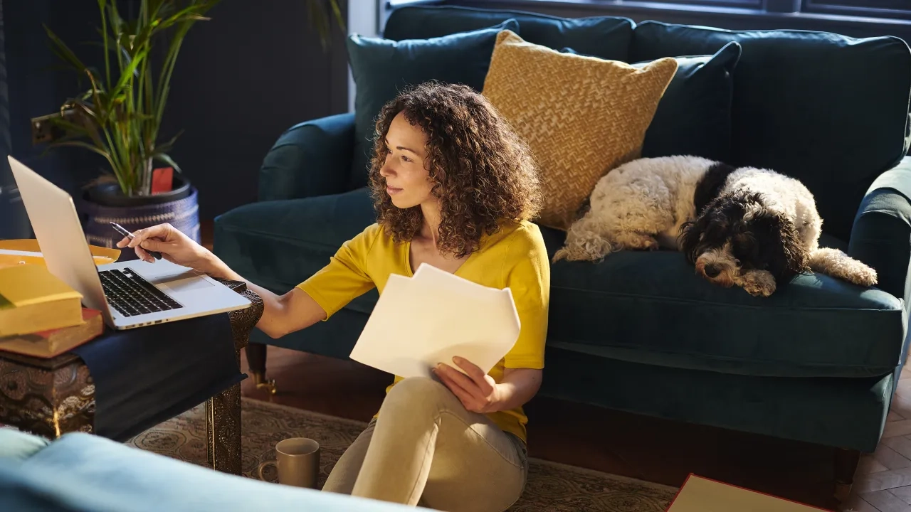 Woman on her laptop reviewing documents with a dog on a nearby couch