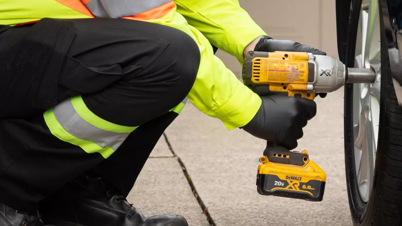 A person wearing a neon yellow jacket and black gloves uses a DeWalt 20V cordless impact wrench to tighten the lug nuts on a car tire. The person is kneeling on a concrete surface beside the dark-colored car.