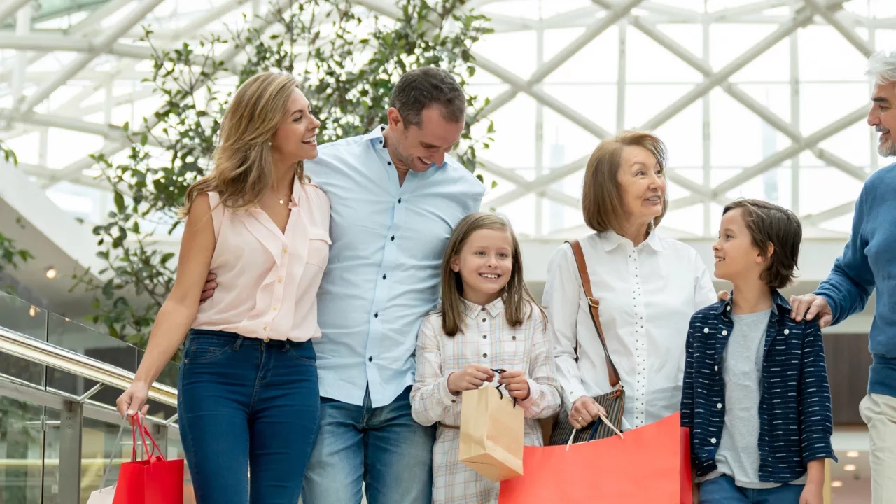 A multi-generational family of six, including children and adults, smiling and carrying shopping bags as they walk through a modern, well-lit shopping mall with a glass ceiling and green plants in the background.