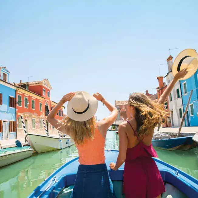 Two women sitting in a boat, enjoying a peaceful canal ride with colorful buildings lining the waterway in the background