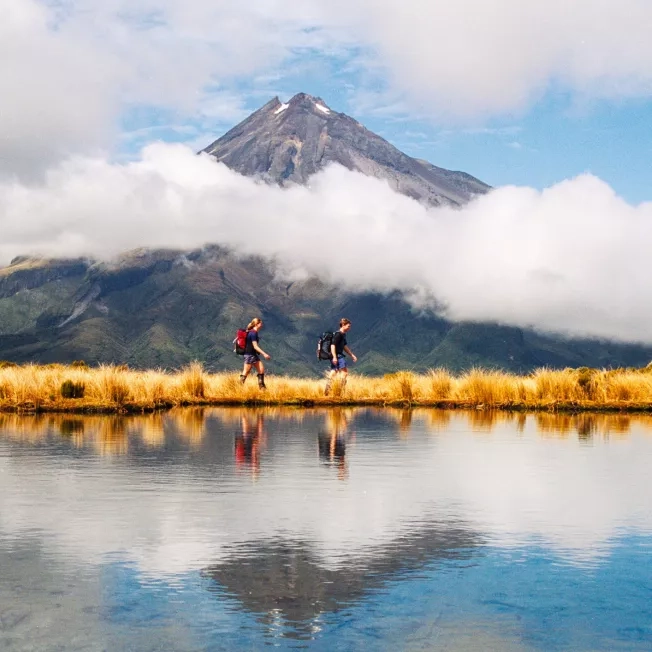 Two hikers walking by Mount Taranaki Egmont, New Zealand, with the reflection along the water.