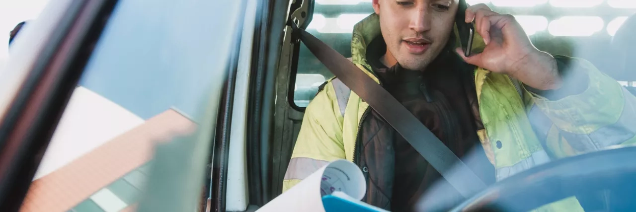 Mid-adult manual worker is sitting in his van, looking through paperwork while speaking to someone on the phone.