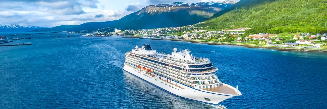 A large white cruise ship sails in blue waters near a coastal town with green hills and mountains in the background. Small houses and buildings line the coast, and the sky is partly cloudy. The picturesque setting shows the natural beauty of the area.