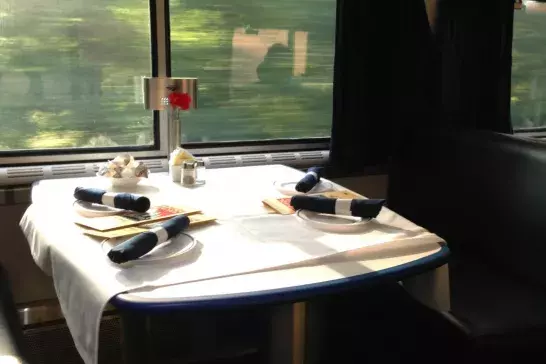 A cozy dining table set inside a train with tableware, menus, and a vase with a red flower, providing a scenic view from the window.