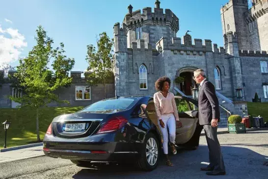 A person stepping out of a black luxury car at the entrance of a grand stone castle, with a suited driver holding the door open