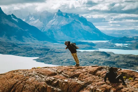 A hiker balances on the edge of a rocky mountain with breathtaking views of the jagged peaks and lakes in the background.