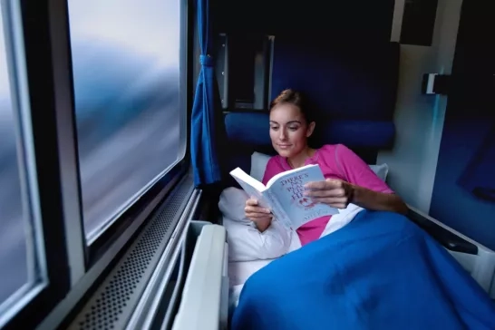  A woman enjoys reading a book while resting in a cozy sleeper car on a train, covered with a blanket, gazing out of the window.