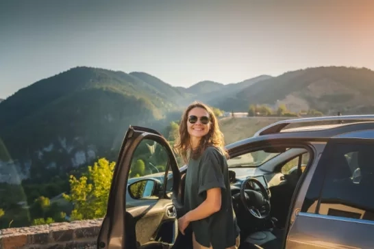 Woman wearing sunglasses standing beside a car rental with mountains in the distance