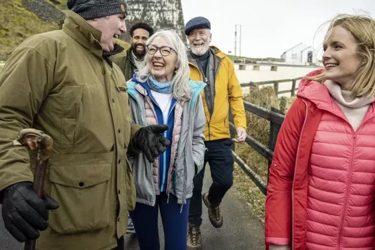 A group of five people dressed warmly, walking and laughing together along a path with green hills in the background.