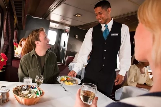 A waiter in a formal vest serves a couple in a train's dining car, with the male passenger receiving a plate of food with a smile.