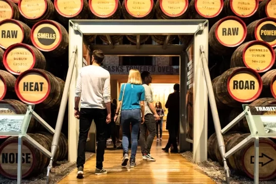 Visitors walking through an exhibit at a brewery, passing by rows of large barrels labeled with different brewing terms such as 'Raw,' 'Smells,' and 'Power