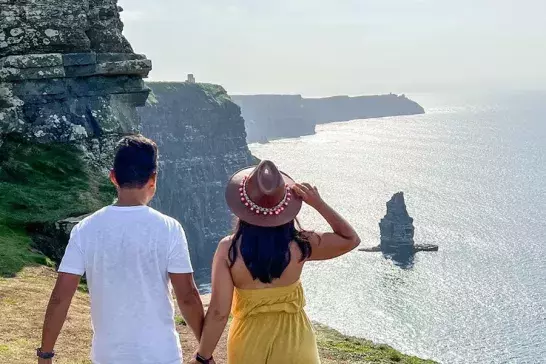 A couple standing hand-in-hand on a cliff, gazing out over a breathtaking ocean view with tall rock formations rising from the water