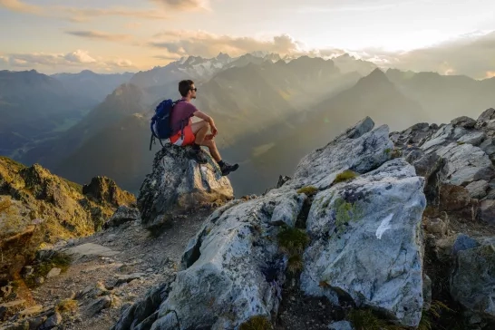 A man sitting on a rock at the summit of a mountain, admiring the scenery after a long hike in Switzerland.