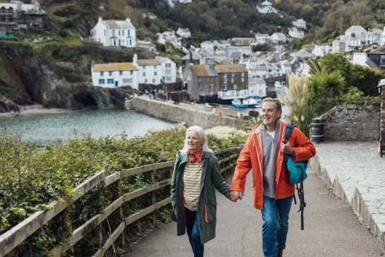 A senior man and his wife holding hands walking up a hill on a footpath looking away from the camera at the view. The fishing village of Polperro is behind them.