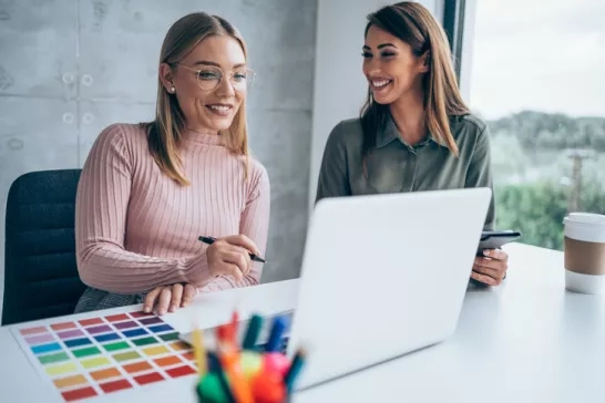 Two women collaborate at a desk, reviewing color options on a laptop, suggesting a creative meeting in progress.