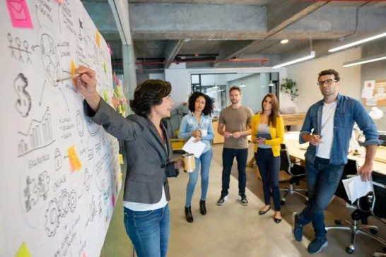 A group of professionals collaborating, with one person writing on a large whiteboard covered in charts, notes, and diagrams.