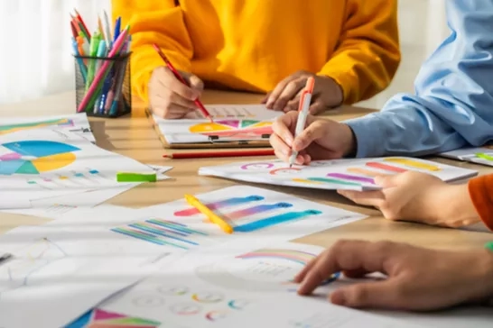 Multiple people working together on a project, surrounded by sheets of paper filled with colorful charts and graphs.