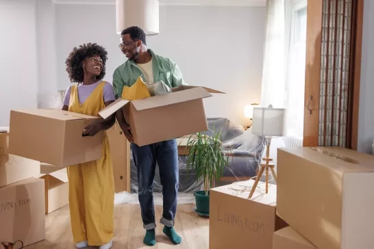 In this heartwarming image, a smiling couple is standing in the sea of boxes in their new living room. The air is filled with excitement as they take the first steps toward making this house a home.