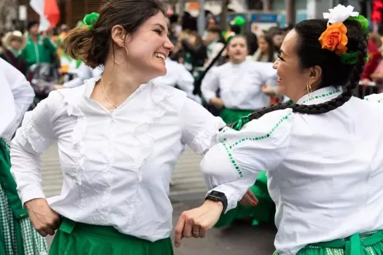 Two women dressed in traditional Irish outfits, smiling and dancing together in celebration, surrounded by other festival participants.