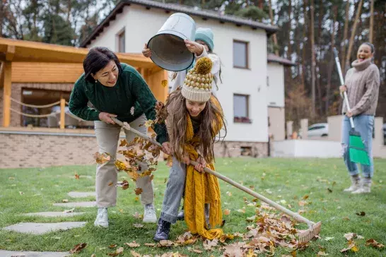 Happy little girls with grandmother picking up leaves and putting them in bucket in garden in autumn Title	