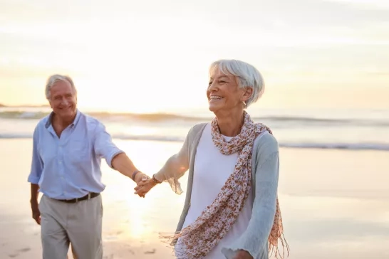 Senior woman laughing while leading her husband along a beach at sunset stock photo