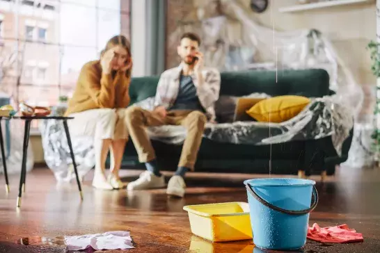 Roof is Leaking or Pipe Rupture at Home: Panicing Couple In Despair Sitting on a Sofa Watching How Water Drips into Buckets in their Living Room. Catastrophe, Distaster and Financial Ruin