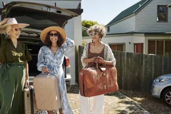 female friends smiling while standing at the trunk of their car full of luggage in a driveway