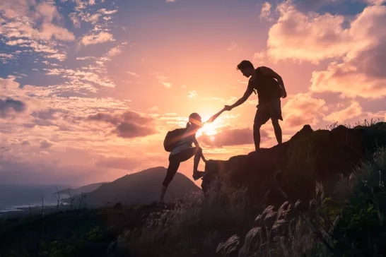 A hiker helping another hiker reach the top of a mountain during sunset.