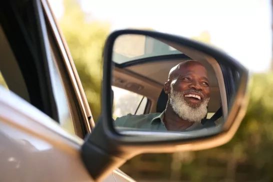 Smiling Senior Male Driver Reflected In Wing Mirror Of Car Enjoying Day Trip Out Driving 