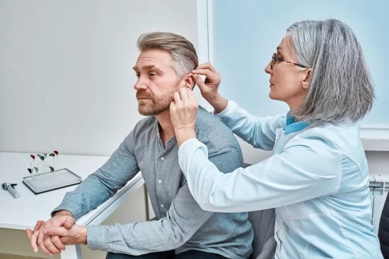 Experienced female doctor fitting Intra-The-Ear hearing aid into patient's ear while consultation in audiology clinic