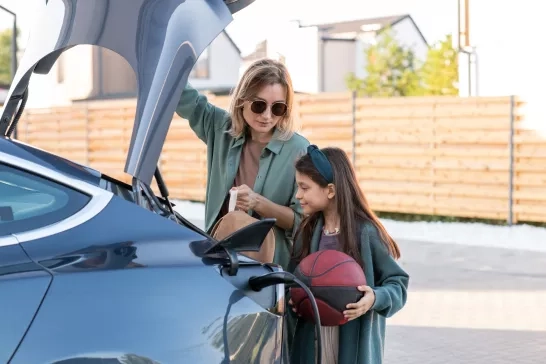 Girl and woman standing by trunk while charging electric car at station