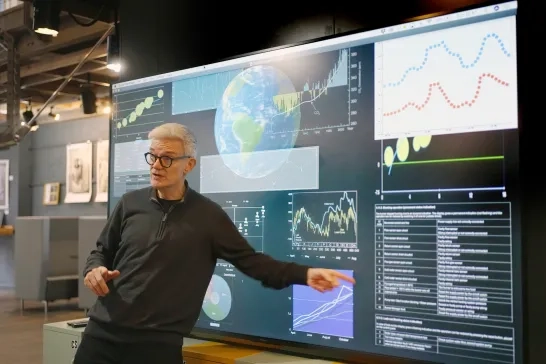 Stock photograph of a mature man giving a lecture in front of a large screen depicting data associated with the planet earth, ie climate change etc .