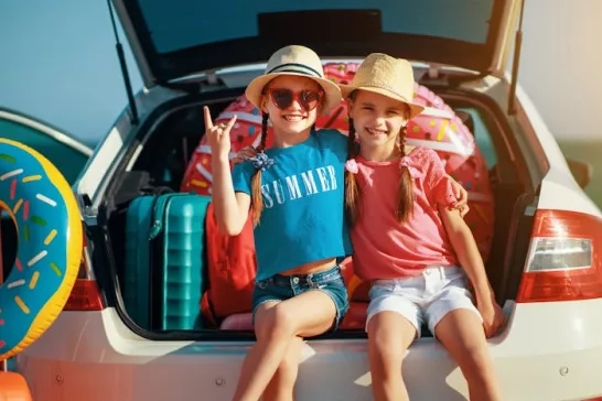 Two kids stting in open trunk of station wagon ready to go on a summer road trip