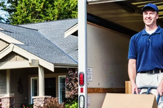 A person wearing a blue shirt and cap smiles while moving a cardboard box using a hand truck. They are standing at the back of a moving truck, which is parked in front of a house with a manicured garden and partial view of a gable roof.