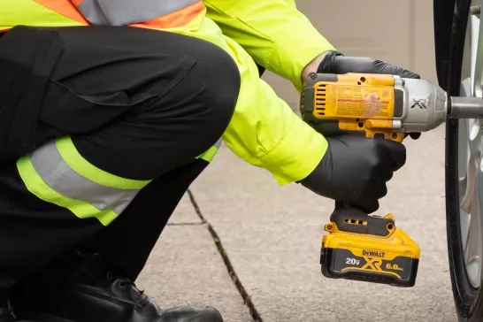 A person wearing a neon yellow jacket and black gloves uses a DeWalt 20V cordless impact wrench to tighten the lug nuts on a car tire. The person is kneeling on a concrete surface beside the dark-colored car.