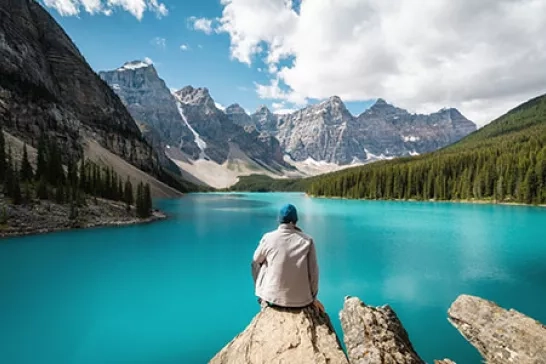 A person in a white jacket and blue hat sits on a rock overlooking a vibrant turquoise lake surrounded by pine trees and towering mountain peaks under a partly cloudy sky.