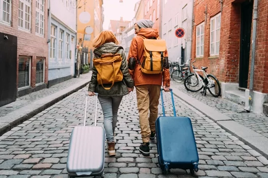 Two people with backpacks and rolling suitcases walk hand-in-hand down a quaint, cobblestone street lined with colorful buildings. Bicycles are parked along the sidewalk, and both individuals are dressed in casual, travel-ready attire.