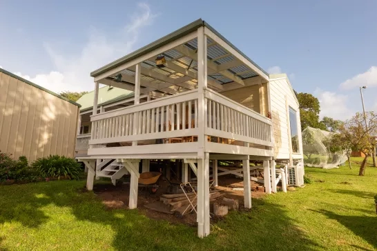 A wooden house on stilts with a covered porch painted white. The house has a metal roof, and various outdoor items such as a wheelbarrow are stored underneath. The surrounding area includes a green lawn, trees, and another structure to the left. The sky is clear and sunny.