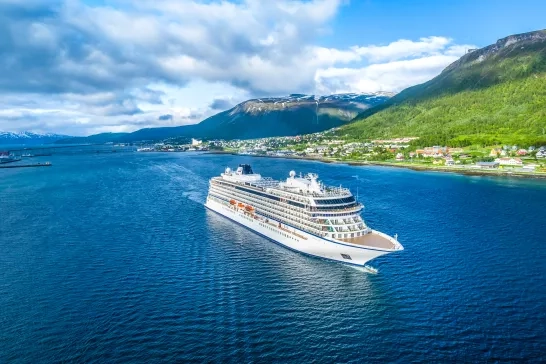 A large white cruise ship sails in blue waters near a coastal town with green hills and mountains in the background. Small houses and buildings line the coast, and the sky is partly cloudy. The picturesque setting shows the natural beauty of the area.