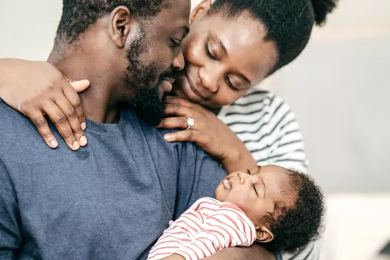 A loving family moment captured as a father holds a sleeping baby in his arms. The mother leans in, embracing both with a tender smile. The baby is dressed in a red and white striped outfit.