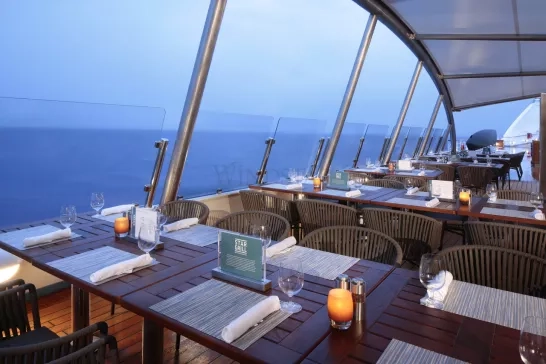 A spacious outdoor dining area on a ship with wooden tables and wicker chairs, each table set with placemats, napkins, wine glasses, and candles. The area is covered by a glass roof and has a clear view of the open ocean in the background.