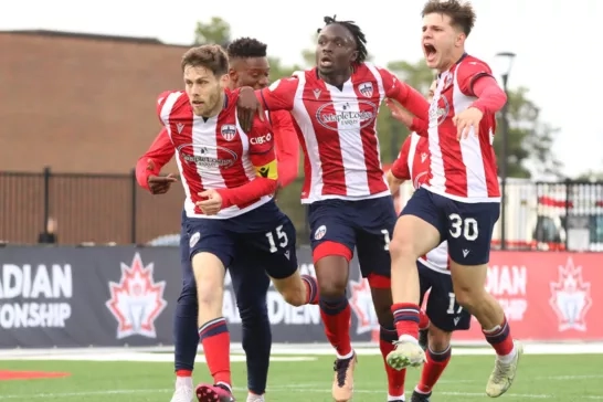 A group of four soccer players in red and white striped jerseys celebrate a goal on the field. Three players have their arms around each other and another player is closely behind. There is a "Canadian Championship" banner in the background.