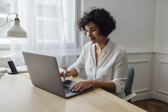 A person wearing a white shirt sits at a wooden desk working on a laptop. The desk holds a smartphone dock, a pen, and some papers. In the background, there is a window with sheer curtains.