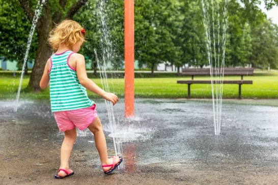 Little smiling child playing at water splash pad fountain in park playground on hot summer day.