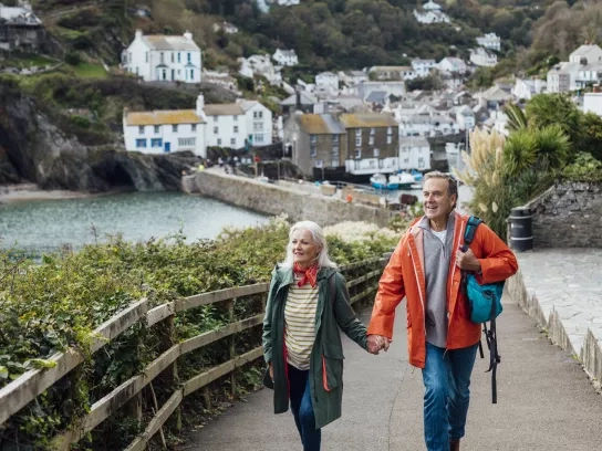 A senior man and his wife holding hands walking up a hill on a footpath looking away from the camera at the view. The fishing village of Polperro is behind them.