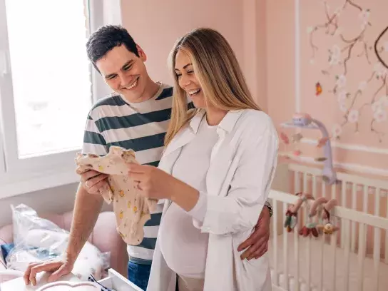 Smiling pregnant woman holding baby outfit with husband in pink nursery