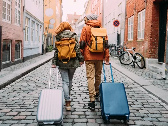 Two people with backpacks and rolling suitcases walk hand-in-hand down a quaint, cobblestone street lined with colorful buildings. Bicycles are parked along the sidewalk, and both individuals are dressed in casual, travel-ready attire.