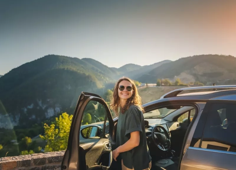 Woman wearing sunglasses standing beside a car rental with mountains in the distance