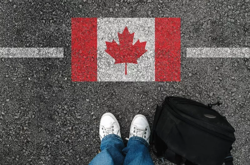 a man with a shoes and backpack is standing on asphalt next to flag of Canada and border